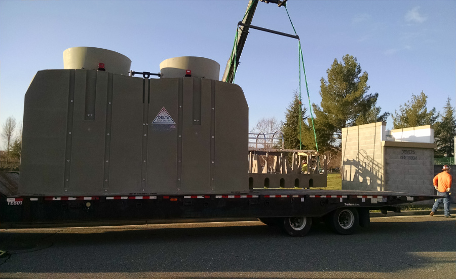 Delta Cooling Towers Beige Twin TM Series on a flatbed of a truck waiting to drive off.  Under the flatbed you can see the black pavement and above it is blue skies. A construction worker is seen in the right side of the image wearing an orange top, jeans and a hard hat. 