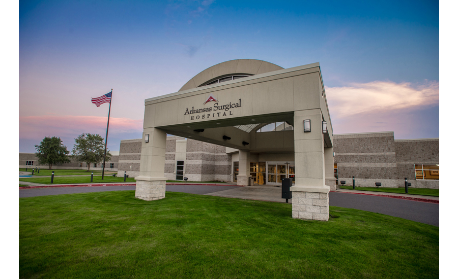 Beige Hospital Building Entrance with green grass on the bottom and a blue sunset with some pink and white clouds. An American flag is blowing in the wind to the right of the hospital. The building is where the anti-microbial cooling towers were installed