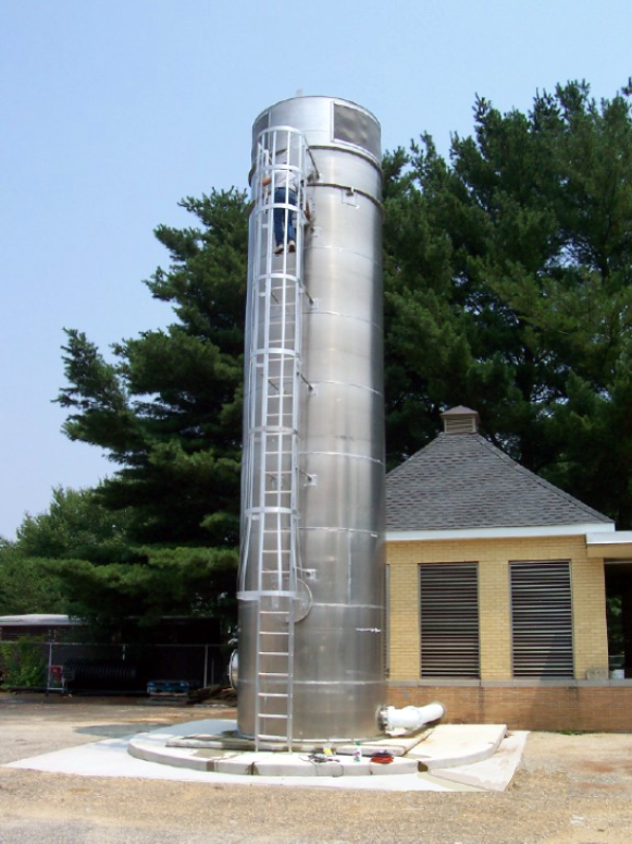 One 7’-6” Diameter x 36’-1” high silver Delta Cooling air stripping tower and (1) 8’-6” Diameter x 36’-1” high air stripping tower to treat potable water.  The tower was installed by a small yellow building with green trees and blue sky in the background.